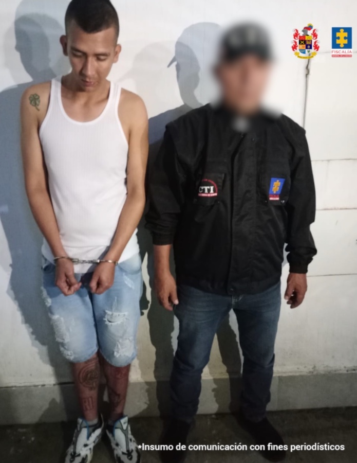 En la fotografía se aprecia de pie, con los brazos adelante y esposado. Viste camiseta esqueleto blanca, bermuda de jean azul desgastado y zapatillas blancas con verde. Al lado izquierdo de él hay un servidor del CTI de la Fiscalía.