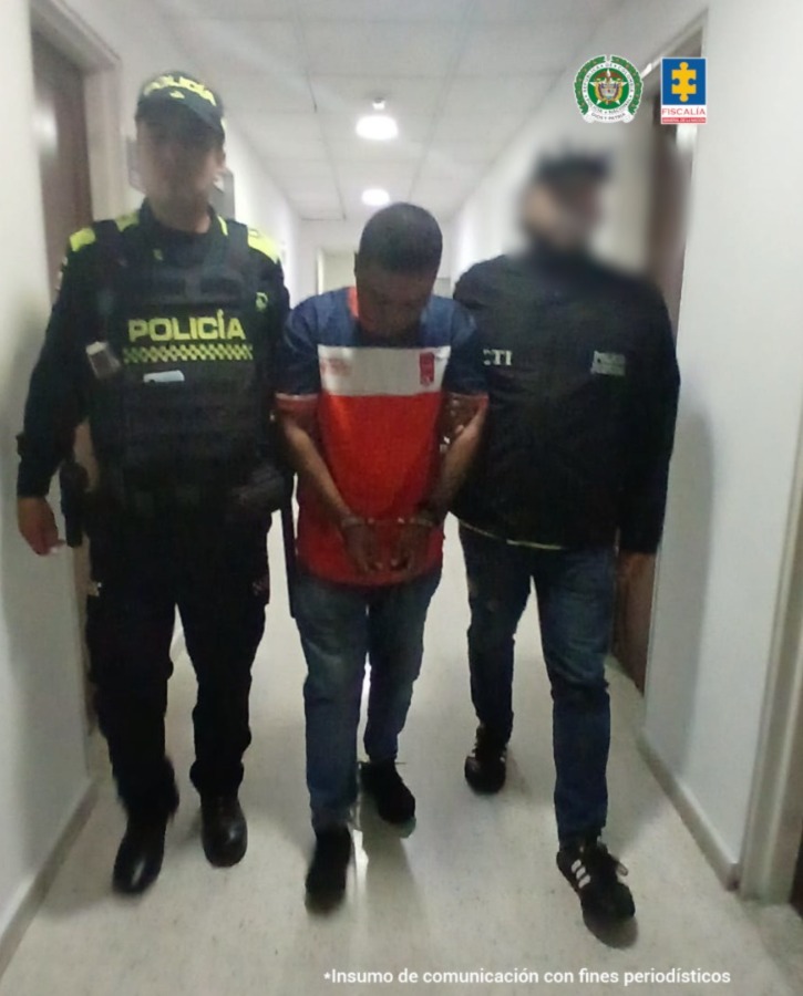 En la fotografía, tomada en un pasillo, en acción de caminata, se aprecia con los brazos adelante y esposado a Jorge Wilmar Olaya Solís. iste camiseta de manga corta, con colores azul, en la parte superior; luego una franja blanca la altura del pecho y del pecho hacia abajo de color rojo. Asimismo, usa jean azul oscuro y zapatillas negras. A su izquierda hay un servidor del CTI de la Fiscalía y a su derecha un uniformado de la Policía Nacional.