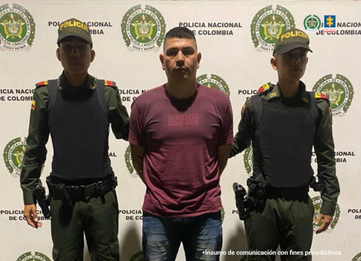 En la fotografía se aprecia de pie y con los brazos atrás a Luis Abrahán Ramírez Mazo. Viste camiseta de color vinotinto, de manga corta y jean azul desgastado. A sus extremos hay uniformados de la Policía Nacional. Detrás de ellos hay un pendón institucional de la Policía.