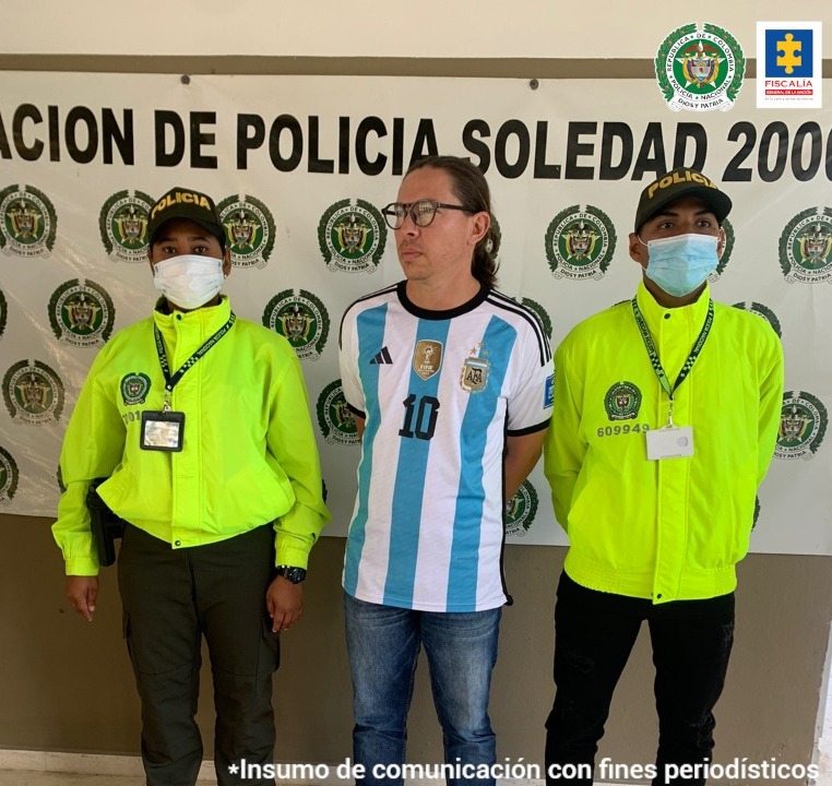 El capturado aparece en el centro de la imagen, con una camiseta de equipo deportivo y gafas. A cada lado tiene a personal de la Policía Nacional.