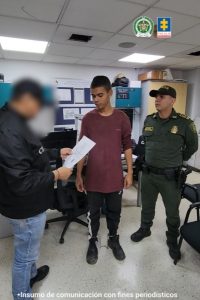 En la fotografía se aprecia de pie, dentro de una oficina a Kevin Eduardo Salcedo Lopera. Viste camiseta de manga corta, de color vinotinto; pantalón de sudadera negro con franja blanca a lo largo, así como zapatillas negras. A la izquierda de él hay un uniformado de la Policía Nacional y a la derecha un servidor del CTI de la Fiscalía quien tiene en sus manos un documento.