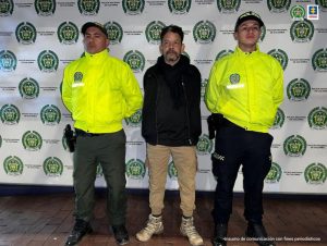 En la fotografía se ven tres personas dos de ellas integrantes de la Policía y uno de ellos el capturado. en la parte posterior se observa un backing con escudos de la Policía Nacional.