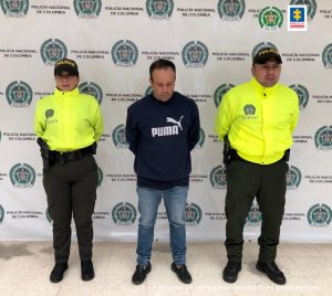 En la fotografía se ven tres personas dos de ellas integrantes de la Policía y uno de ellos el capturado. en la parte posterior se observa un backing con escudos de la Policía Nacional. 