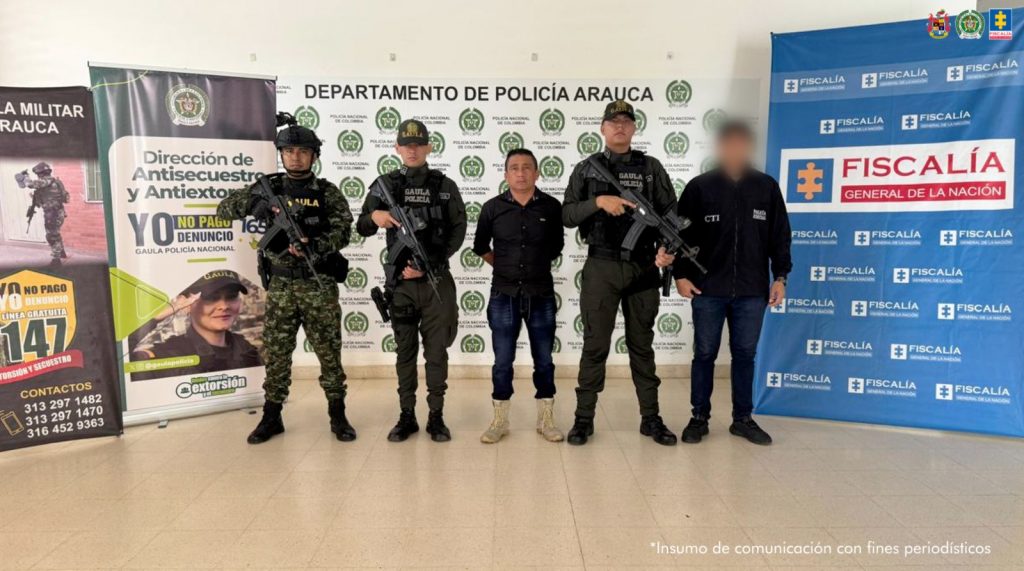 Se visualiza al capturado junto a uniformados del Gaula Policia y Militar de Arauca e investigador del CTI. Detrás los banners que identifican a la Fiscalía General de la Nación y al Ejército y Policía Nacional.
