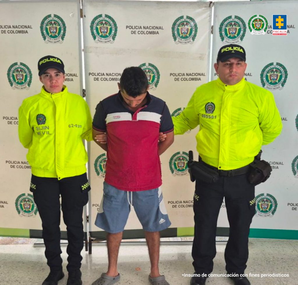 capturado de frente custodiado por dos policías. Detrás banner de la Policía Nacional.