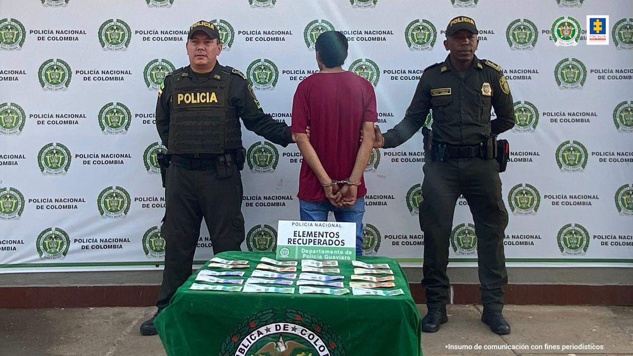 Está el detenido de espaldas esposado custodiado por dos policías uniformados. Detrás banner de la Policía Nacional.