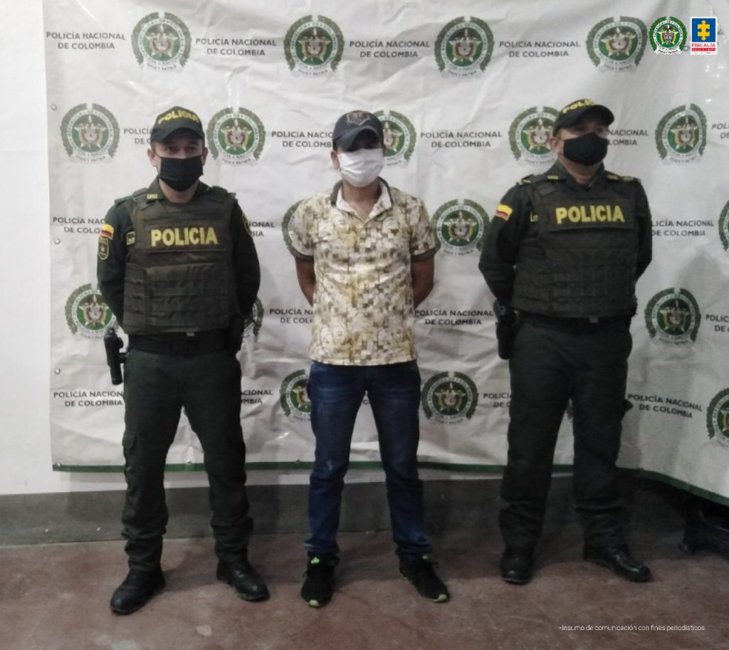 en la imagen se ve una persona detenida bajo custodia de dos integrantes de la Policía Nacional. Detrás suyo un backing de la institución.