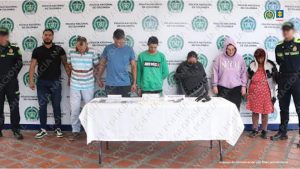 Se observan siete personas capturadas, en fila y de frente. A cada costado un funcionario de Policía nacional. Atrás un pendón de Policía nacional. Adelante una mesa con material incautado durante allanamientos