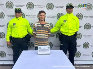 Se observa al capturado de frente y custodiado por dos funcionarios de la Policía Nacional que se ubican uno a cada lado. Atrás , un pendón de Policía Nacional. Adelante una mesa en donde está un aviso que dice: Captura Orden Judicial