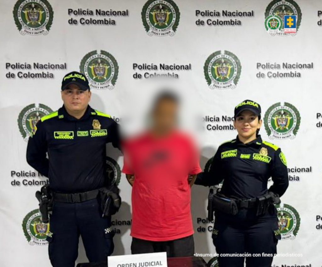 En la fotografía aparece de pie y con los brazos atrás el hoy imputado. Viste con camiseta de color rojo, con estampado de color negro en el pecho y pantalón negro. En los extremos hay uniformados de la Policía Nacional. Delante de ellos hay una superficie de color marrón y sobre ella hay un letrero de color blanco que dice: ‘Orden Judicial’. Detrás de ellos hay un pendón institucional de la Policía.