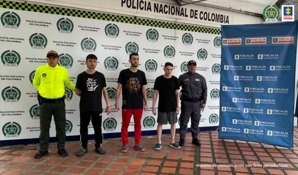 Se observan tres detenidos, a un costado un funcionario del CTI y al otro uno de Policía Nacional.  Atrás un pendón de la Fiscalía General de la Nación y otro de Policía Nacional.