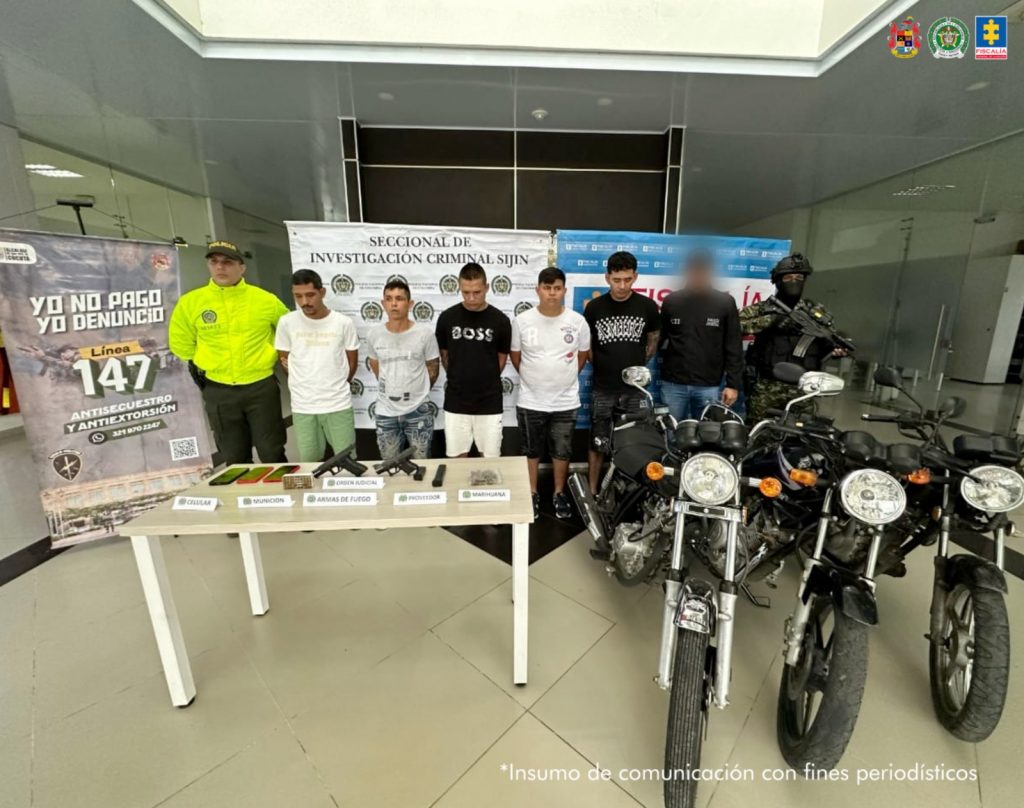 Se visualiza a los capturados junto a un investigador del CTI y uniformados del Ejército y Policía Nacional. Detrás de ellos los banners que identifican a la Fiscalía General de la Nacional, Ejército y Policía Nacional.