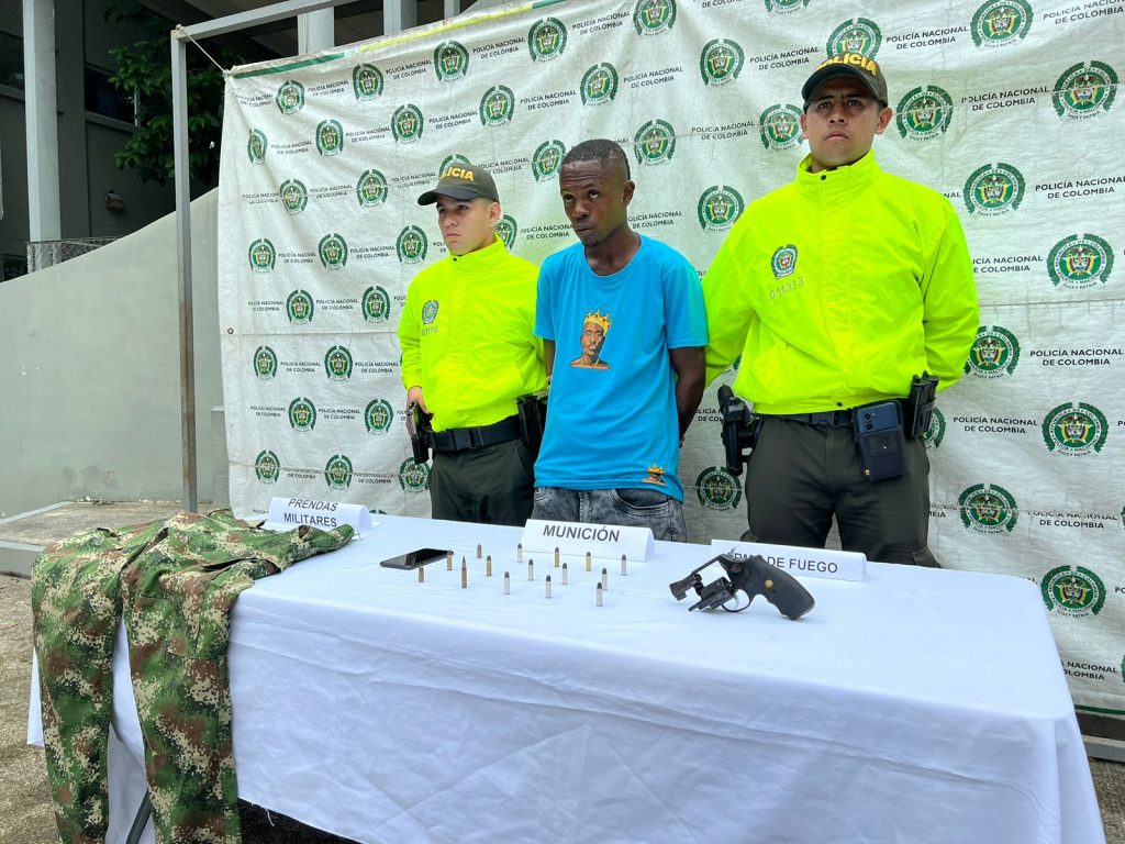 se observa al detenido en el centro y a cada costado un funcionario de Policía nacional. Atrás un pendón de Policía Nacional y al frente una mesa con material incautado en el operativo de allanamiento y registro