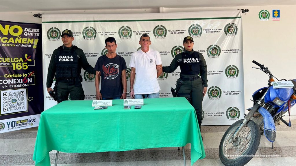 Los capturados se encuentran en custodia por dos uniformados del Gaula de la Policía Nacional. Detrás hay un backing de la Policía. En la foto se observa parte del material incautado (moto, celular y dinero en efectivo)