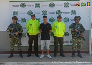 capturado de frente custodiado por dos policías y dos militares. Detrás banner de la Policía Nacional.