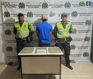 En la fotografía se aprecia de espaldas a la cámara y de pie a Gionaiker Alejandro Bracamonte Bárcenas. Viste camisa de manga corta color azul petróleo, pantalón oscuro y gorra color caqui. A los extremos de él hay uniformados de la Policía Nacional. Frente a ellos hay una mesa y sobre ella varios paquetes en bolsas plásticas con contenido verdoso. En la parte posterior hay un pendón institucional.