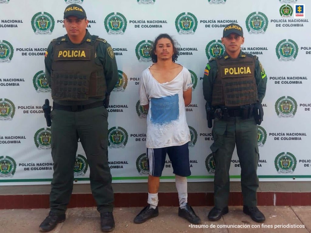 En la foto se ve un hombre parado de frente con cabello desordenado brazos atrás, en camiseta, pantaloneta, medias y zapatos sin cordones custodiado por dos uniformados de la Policía Nacional. Detrás el banner de la Policía.