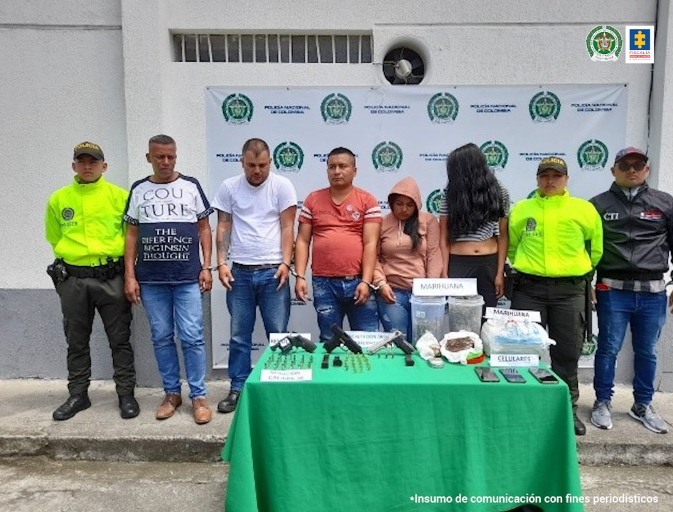 En la foto aparecen los cinco presuntos integrantes del grupo delictivo ‘El Indio’. Tres hombres y dos mujeres. Al lado derecho de ellos hay un uniformado de la Policía y al izquierdo un servidor del CTI de la Fiscalía y otro uniformado de la Policía. Delante de ellos hay una mesa con mantel verde y sobre ella hay tres armas de fuego, munición, estupefacientes y 3 teléfonos celulares. Detrás de ellos hay un pendón institucional de la Policía Nacional.
