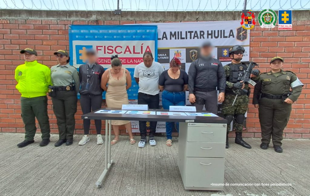 Los capturados se encuentran de frente y custodiados por servidores del Cuerpo Técnico de Investigación CTI, Ejército y Policía Nacional. En la foto se observa un backing de la Fiscalía y Ejército. También se observa las tarjetas que se incautaron durante el allanamiento.