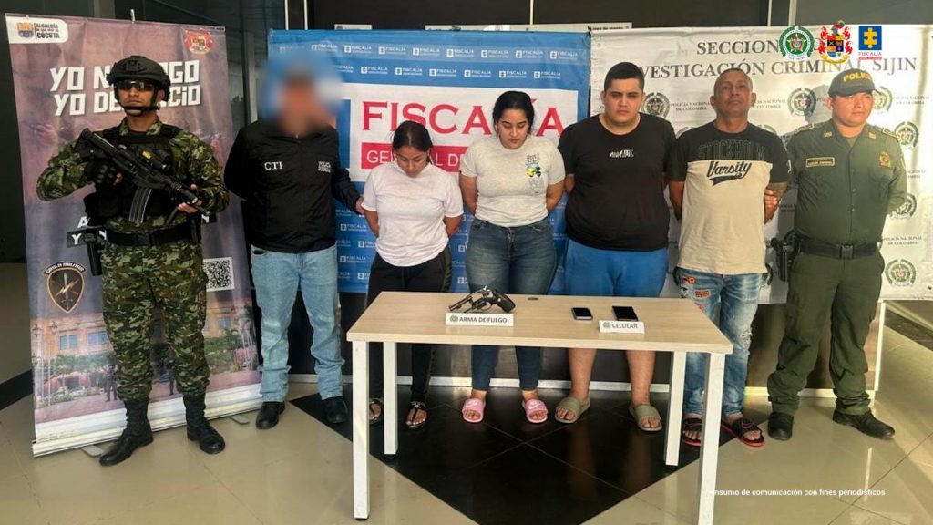Se visualiza a los capturados junto a investigador del CTI , y uniformados del Ejército y Policía Nacional. Frente a ellos los elementos incautados: un arma de fuego y dos teléfonos celulares. Detrás de ellos los banners institucionales de la Fiscalía General de la Nación, Ejército y Policía Nacional.