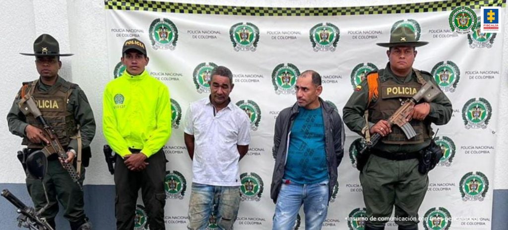 En la foto aparecen, de pie, con los brazos atrás, dos de los tres presuntos integrantes de Los Satiros. Uno viste camiseta blanca de manga corta jean azul claro desgastado. La otra persona viste con chaqueta oscura y camiseta verde con estampado, sí como jean azul. A lado y lado de ellos hay uniformados de la Policía Nacional. Detrás de ellos hay un pendón institucional de la Policía.