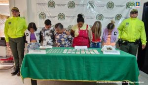 Los capturados se encuentran de frente y custodiados por dos uniformados de la Policía Nacional. Detrás hay un backing de la Policía Nacional. En la foto se observa una mesa con parte del material incautado como marihuana, dinero en efectivo y gramera.
