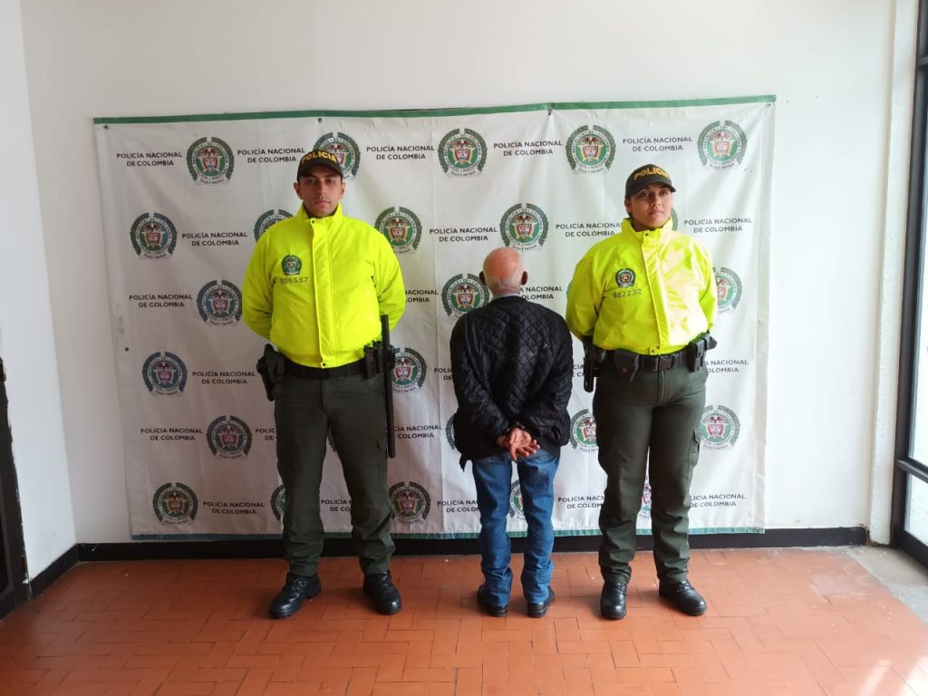 Se observa al detenido de espaldas y a cada costado un funcionario de Policía Nacional. Al fondo pendón de Policía Nacional