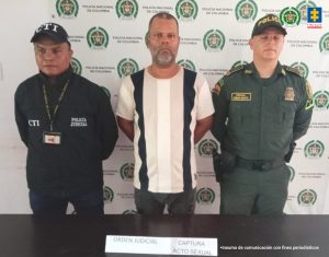 En la fotografía aparece de pie y con los brazos atrás a Gustavo Adolfo Vásquez Marmolejo. Viste con camiseta de manga corta con franjas verticales negras; pantalón oscuro. Al lado derecho de él hay un servidor del CTI de la Fiscalía y al lado izquierdo un uniformado de la Policía Nacional. Delante de ellos hay una superficie color marrón y sobre ella hay un letrero que dice: ‘Captura orden judicial. Acto sexual’. Detrás de ellos hay un pendón institucional de la Policía.