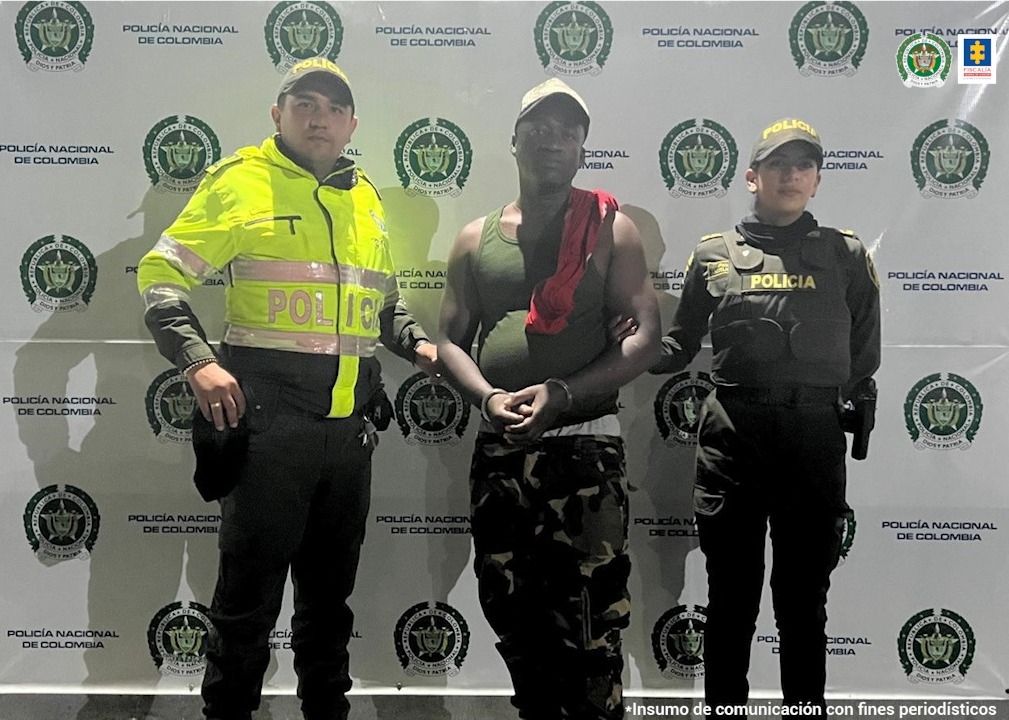 En la foto aparece, de pie Gustavo Angulo Quiñones, con los brazos adelante y esposas en sus muñecas. Viste camiseta esqueleto verde, pantalón tipo camuflado y gorra. Asimismo, tiene una tela roja sobre su hombro izquierdo. A su derecha e izquierda hay uniformados de la Policía Nacional. Detrás de ellos hay un pendón institucional de la Policía.