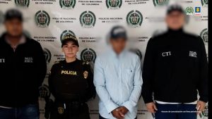 En la fotografía se aprecia de pie y con los brazos adelante a Gustavo Bedoya Saavedra. Viste camisa de manga larga azul claro, pantalón negro, gorra negra con estampado impreso de color blanco al frente. A sus extremos hay servidores del CTI de la Fiscalía y uniformados de la Policía Nacional. Detrás de ellos hay un pendón institucional de la Policía.
