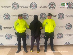 Se observa al capturado de espaldas y a cada costado un funcionario de Policía Nacional. Atrás un pendón de Policía Nacional