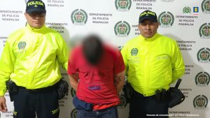  Se observa al capturado de frente. A cada costado un funcionario de la Policía Nacional. Atrás un pendón de Policía Nacional.