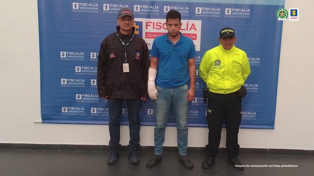 se observa al detenido y a un costado un funcionario del CTI y al otro uno de Policía nacional. Al fondo un pendón de la Fiscalía General de la Nación