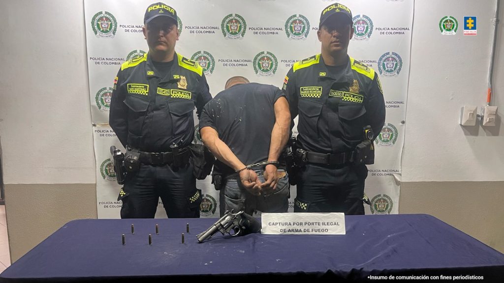 Se observa al capturado de espaldas, esposado.  A cada costado un funcionario de Policía Nacional, atrás un pendón de Policía Nacional y adelante una mesa con el material incautadoPIE DE FOTO: Hernando de Jesús Otálvaro Acevedo habría sido sorprendido portando un revólver calibre 38, con munición.  