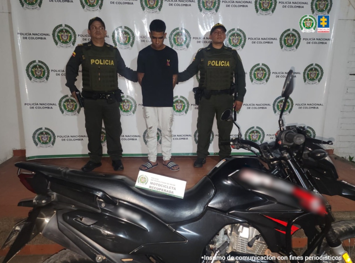 El capturado se encuentra de frente y custodiado por dos uniformados de la Policía Nacional. En la foto se ve de fondo un backing de la Policía Nacional y la motocicleta que se había hurtado.
