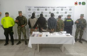 En la foto aparecen 4 hombres de espaldas custodiados por dos Policías y dos soldados del Ejército Nacional, adelante de ellos hay una mesa con dos armas de fuego, municiones y dinero en efectivo