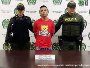 En la foto aparece de pie, los brazos atrás Hámilton Andrés Agudelo, alias ‘Cholo’. El hombre viste camiseta roja con el estampado de un oso amarillo con un letrero que dice ‘Moschino’. A lado y lado de él hay uniformados de la Policía Nacional. Delante de ellos hay una mesa y sobre ella hay un letrero que dice ‘Captura por orden judicial’. Detrás de ellos hay un pendón institucional de la Policía.