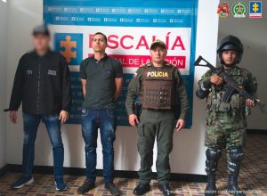 El imputado se encuentran en custodia por un servidor del Cuerpo Técnico de Investigación (CTI), un uniformado de la Policía y el Ejército Nacional. Detrás de ellos hay un backing de la Fiscalía. 