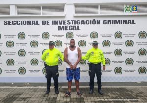 En la fotografía se aprecia de pie y con los brazos adelante y esposado a Harvi Alejandro García Rúa. Viste con camisa esqueleto blanca, bermudas de color azul estampado y pantuflas azules con blanco. Usa barba tipo ‘chivera’. A sus extremos hay uniformados de la Policía Nacional. Detrás de ellos hay un pendón institucional de la Policía.