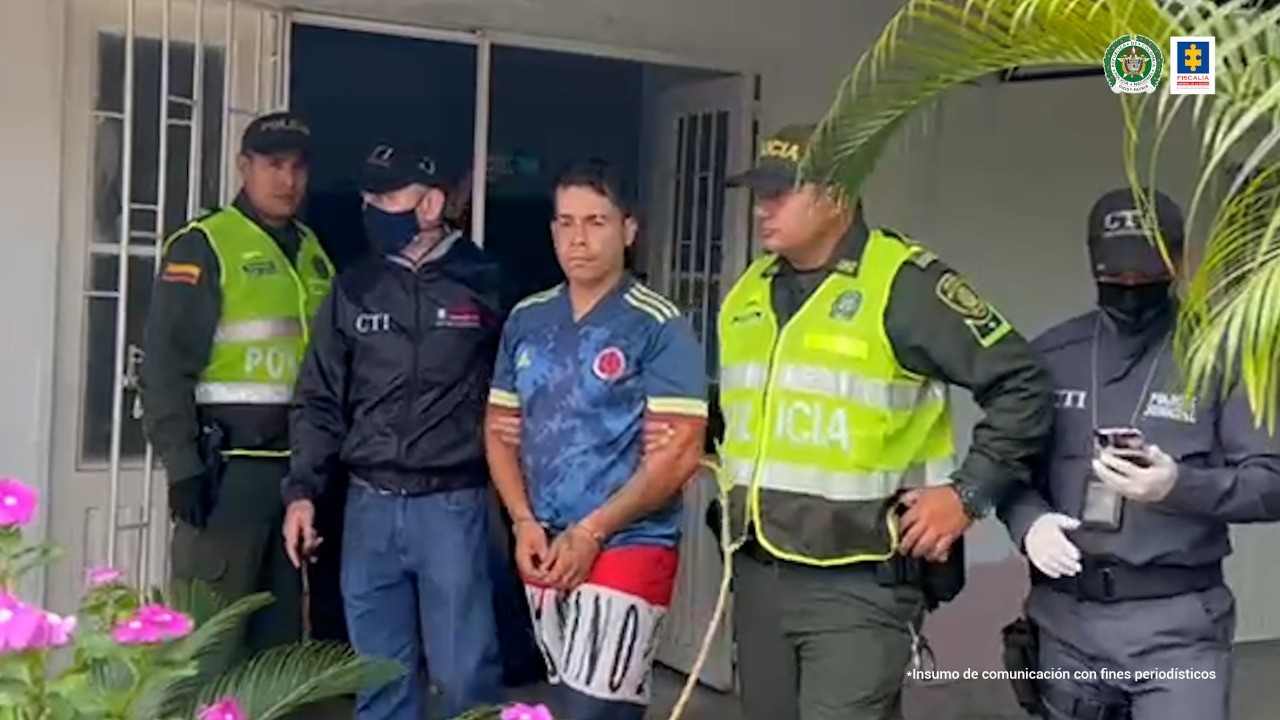 En la foto se ven llevando al capturado esposado, vestido con camiseta y pantaloneta, a un investigador del CTI de la Fiscalía y un Policía uniformados acompañados de dos unidades más de cada Institución saliendo de la vivienda donde se llevó a cabo la captura. Detrás se ce una puerta abierta, plantas y flores.