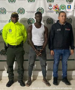 En la foto aparece Héctor Danilo Caicedo Cuero, alias ‘Tyson’, con los brazos adelante y esposado. Vestido con camiseta esqueleto blanca, pantalón oscuro y pantuflas. Al lado derecho de él hay un uniformado de la Policía Nacional y a la izquierda un servidor del CTI de la Fiscalía. Detrás de ellos hay un pendón institucional de la Policía Nacional. 