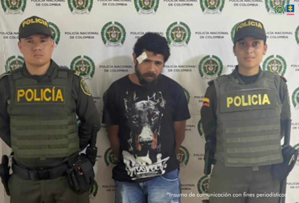 En la fotografía se aprecia de pie y con los brazos atrás. Usa barba y bigote poblados. Viste camiseta de color negro con estampado en la parte frontal; así como jean azul. A sus extremos hay uniformados de la Policía Nacional. Detrás de ellos hay un pendón institucional de la Policía.
