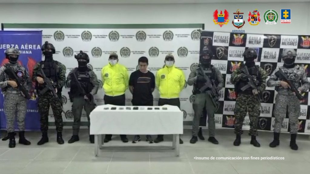 Se visualiza al capturado junto a uniformados del Gaula Militar, Policía Nacional, Armada Nacional y Fuerza Aeroespacial. Frente a ellos una mesa con los nueve celulares incautados. Detrás los banners institucionales de Ejército, Policía y Armada Nacional,  y Fuerza Aérea Colombiana