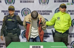 capturado custodiado por dos policías. Detrás está un banner de la Policía Nacional.
