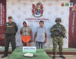 capturada esposados, custodiados por un militar y un policía uniformados, delante de ellos en una mesa droga incautada y detrás banner de Ejército Nacional