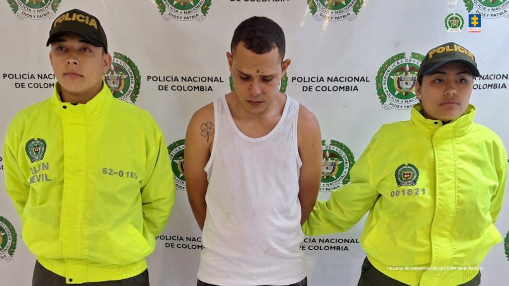capturado de frente custodiado por dos policías. Detrás banner de la Policía Nacional.