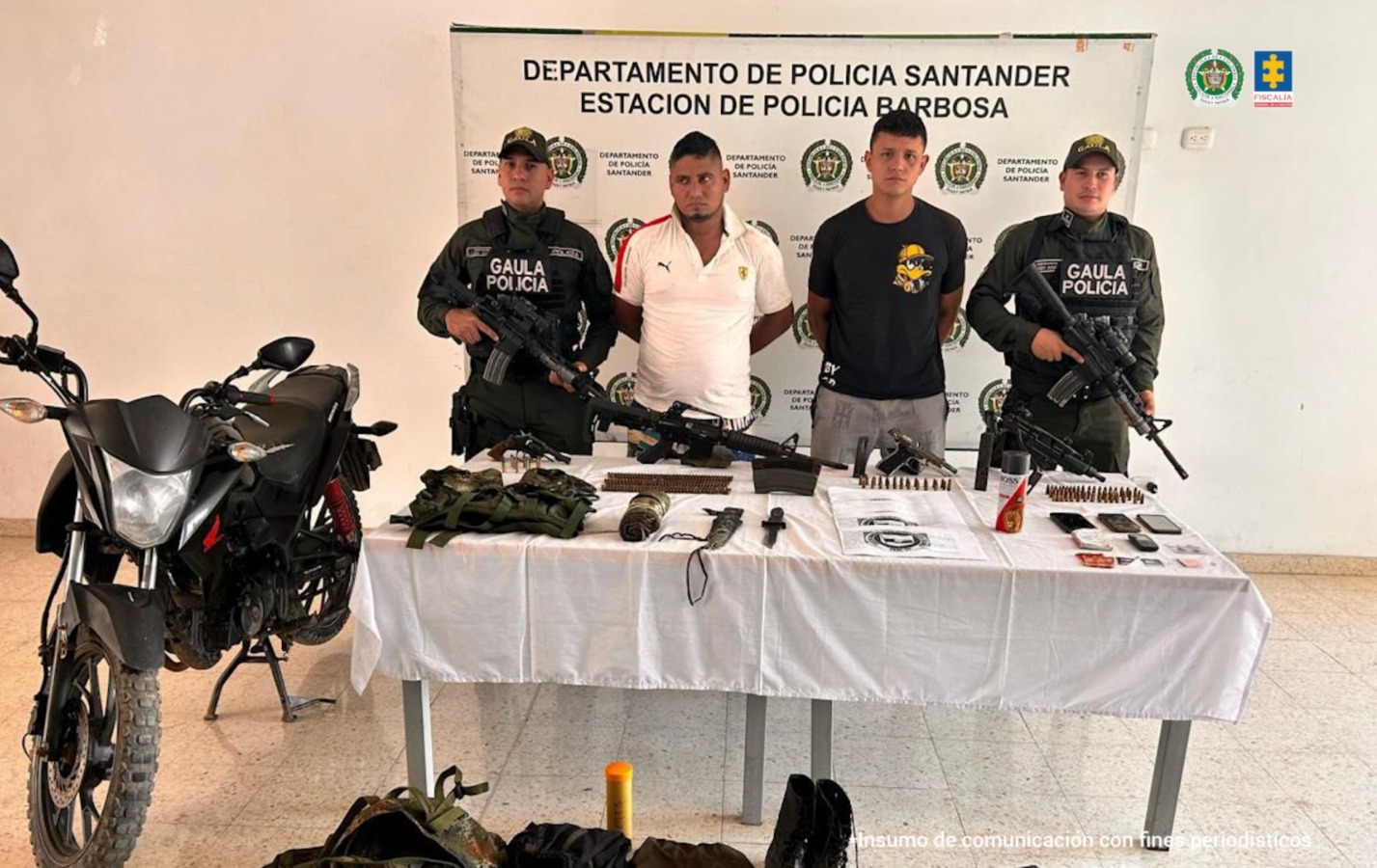 Los hombres capturados aparecen detrás de una mesa, esposados con las manos atrás. Están rodeados de miembros del Gaula Policía. Frente a ellos hay u na mesa con varios elementos y armas, y una motocicleta en el lado izquierdo. 