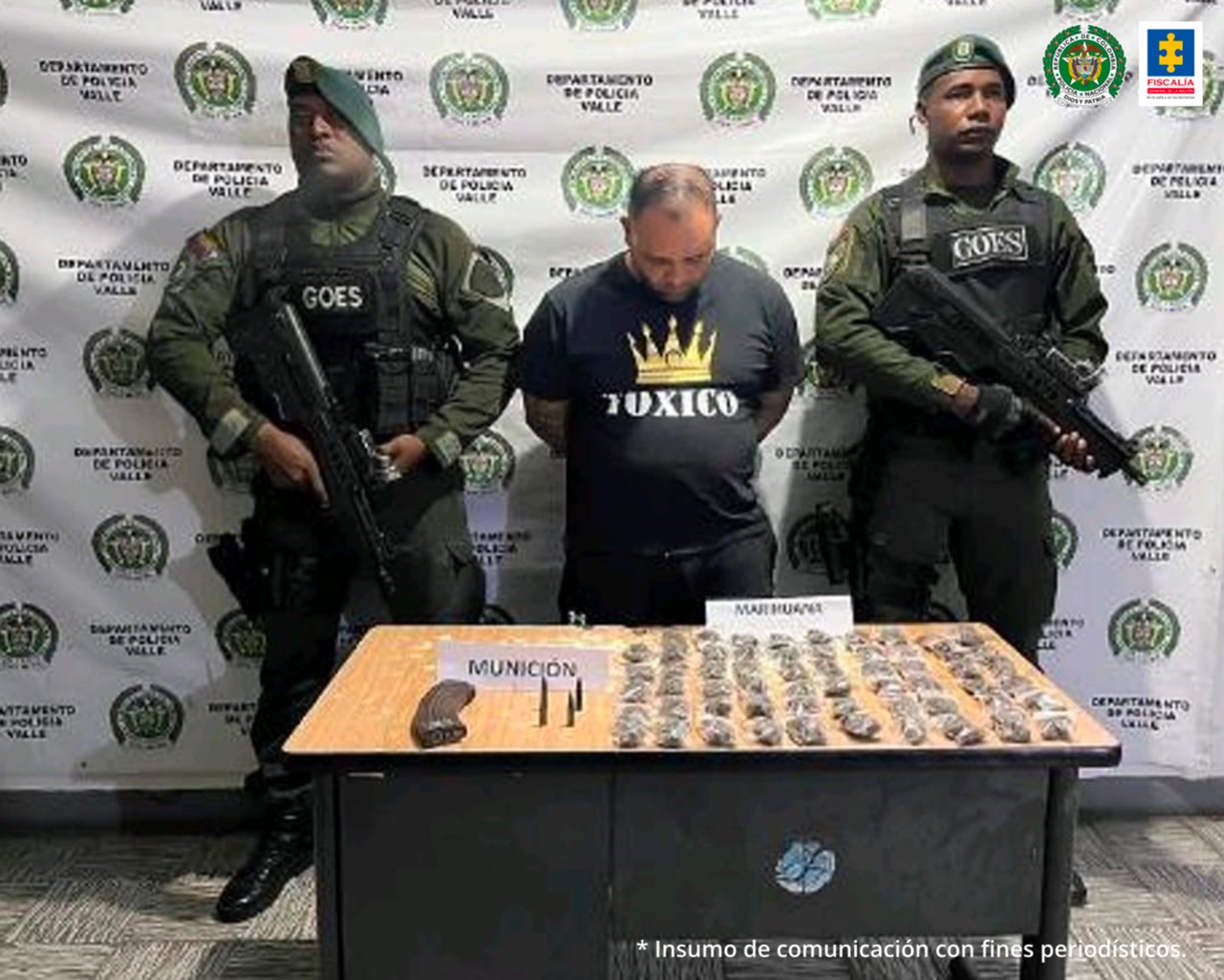En la foto aparece el judicializado con los brazos atrás, vestido con una camiseta negra con estampado de una corona amarilla y letras blancas. A lado y lado de él hay dos uniformados de la Policía Nacional. Delante de ellos hay una mesa con dos letreros que dicen marihuana y munición. Detrás del judicializado y los policiales hay un pendón institucional de la Policía Nacional.