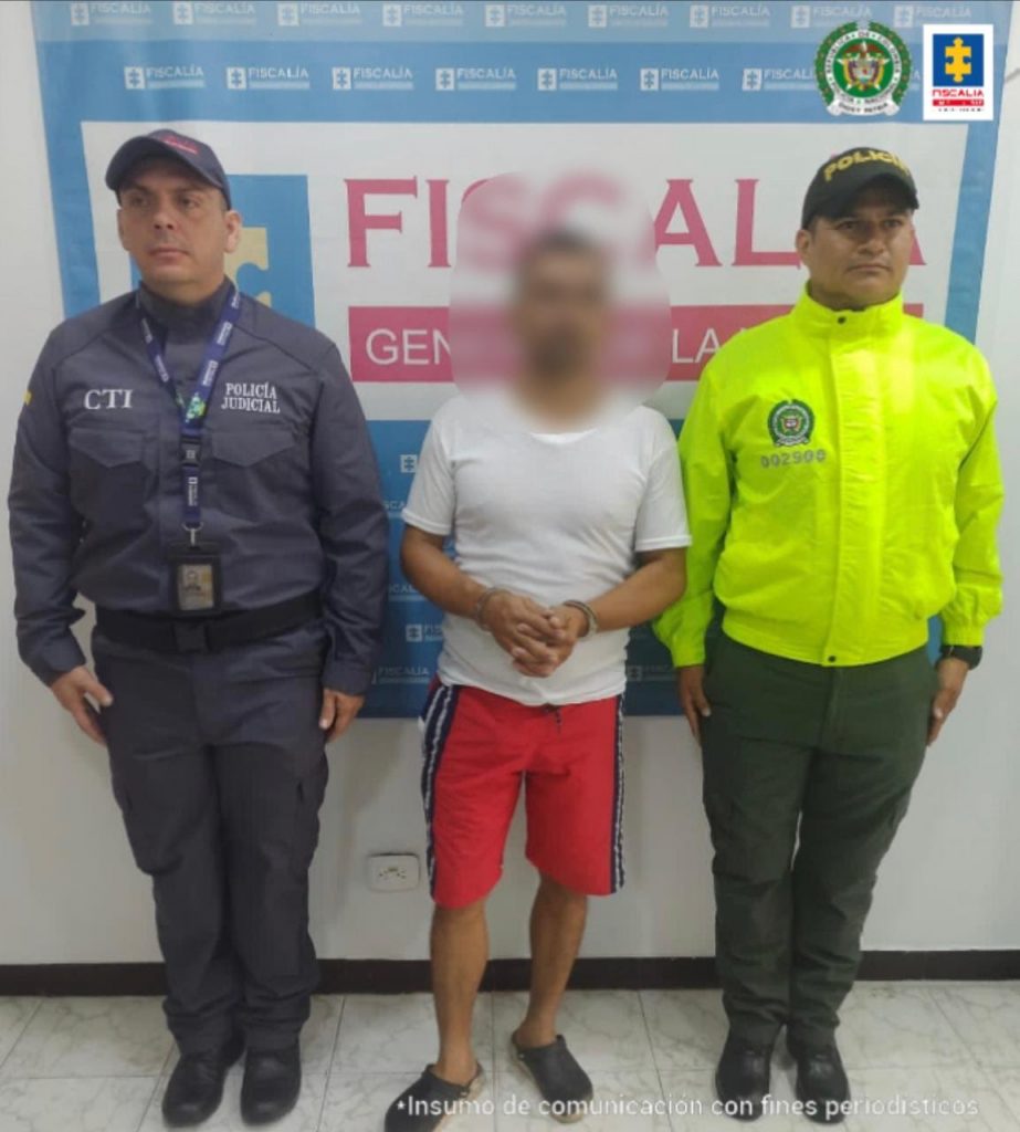 está el detenido de frente esposado y custodiado por investigadores del CTI de la Fiscalía y de la Policía Nacional. Detrás banner de la Fiscalía.