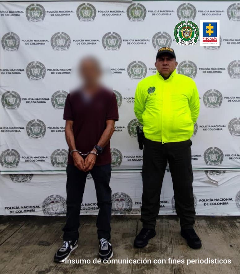El capturado se encuentra esposado y es custodiado por un agente de la Policía Nacional. En la parte de atrás está en pendón institucional.